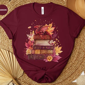 May include: Maroon t-shirt featuring a watercolor illustration of a red dragon perched atop a stack of books. The design includes autumn leaves and flowers in shades of yellow, orange, and pink. The shirt has a crew neck and short sleeves.