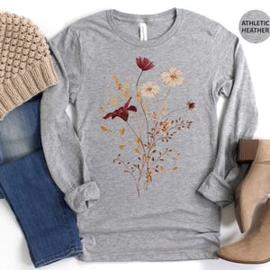 Op de afbeelding: Grijs gem&ecirc;leerd shirt met lange mouwen met een bloemenontwerp in bordeauxrood en cr&egrave;me. De afbeelding bevat een beige gebreide muts, een blauwe jeans en beige enkellaarsjes. De tekst "ATHLETIC HEATHER" staat in een cirkel.