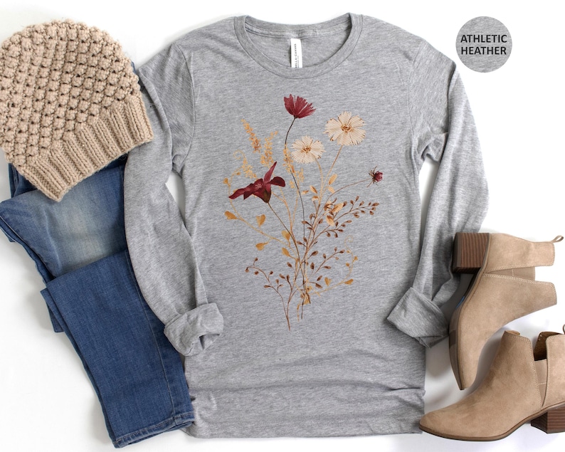 Op de afbeelding: Grijs gem&ecirc;leerd shirt met lange mouwen met een bloemenontwerp in bordeauxrood en cr&egrave;me. De afbeelding bevat een beige gebreide muts, een blauwe jeans en beige enkellaarsjes. De tekst "ATHLETIC HEATHER" staat in een cirkel.