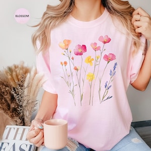 K&ouml;nnte beinhalten: Ein pinkfarbenes T-Shirt mit einem Aquarell-Blumenmuster mit rosa, gelben und violetten Blumen. Das Shirt hat einen Rundhalsausschnitt und kurze &Auml;rmel.
