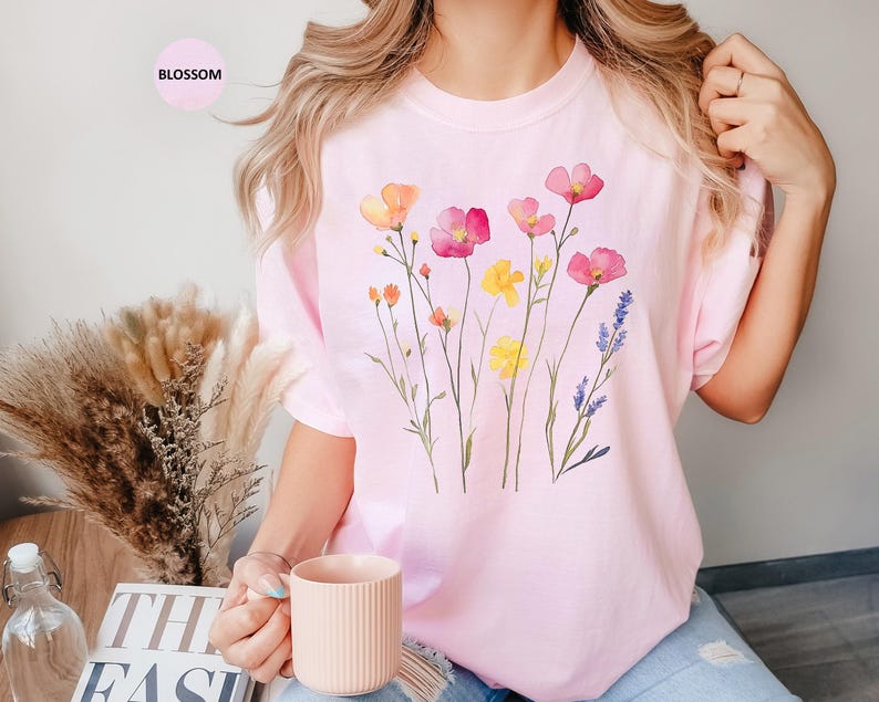 K&ouml;nnte beinhalten: Ein pinkfarbenes T-Shirt mit einem Aquarell-Blumenmuster mit rosa, gelben und violetten Blumen. Das Shirt hat einen Rundhalsausschnitt und kurze &Auml;rmel.