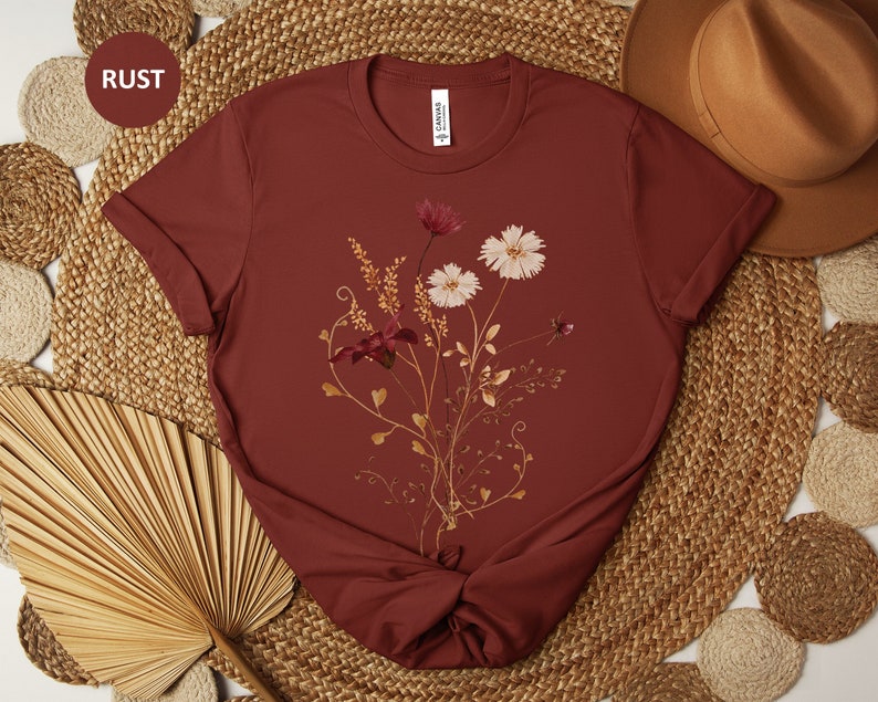 May include: A rust coloured t-shirt with a floral design featuring dried flowers in shades of brown, white, and pink. The shirt has a crew neck and short sleeves.