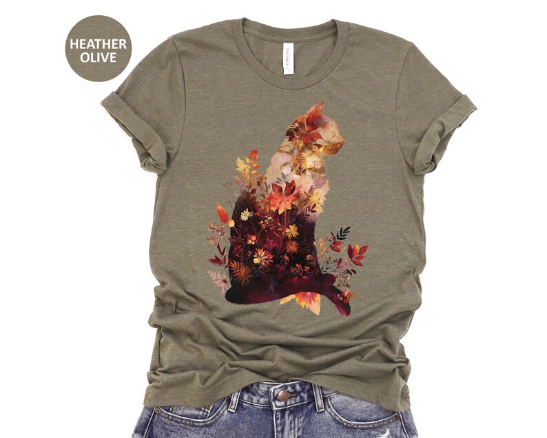 May include: Heather olive t-shirt featuring a cat silhouette filled with autumn leaves in shades of red, orange, and yellow. The shirt is made of a soft material and has a classic crew neck design. The text "HEATHER OLIVE" is in a circle.