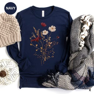 Op de afbeelding: Marineblauwe longsleeve shirt met een bloemenontwerp. Een beige gebreide beanie, een pompoen met patroon en een sjaal met patroon met katoenen accenten staan ook in de afbeelding. Het woord "NAVY" staat in een marineblauwe cirkel.