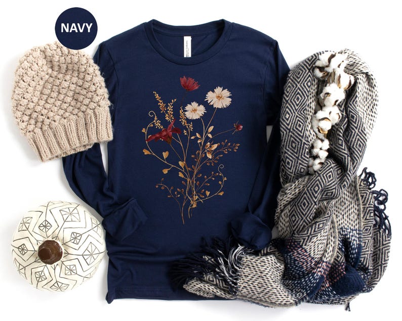 Op de afbeelding: Marineblauwe longsleeve shirt met een bloemenontwerp. Een beige gebreide beanie, een pompoen met patroon en een sjaal met patroon met katoenen accenten staan ook in de afbeelding. Het woord "NAVY" staat in een marineblauwe cirkel.