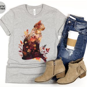 May include: A light gray t-shirt with a cat silhouette filled with autumn leaves and flowers. The shirt is paired with blue jeans, tan ankle boots, and a coffee cup. The text "ATHLETIC HEATHER" is printed on a gray circle.