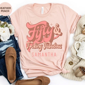 May include: A heather peach t-shirt with the text "Fifty & F*cking Fabulous" in a retro font with a pink and brown color scheme. The name "Samantha" is printed below the text.