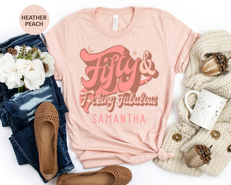 May include: A heather peach t-shirt with the text "Fifty & F*cking Fabulous" in a retro font with a pink and brown color scheme. The name "Samantha" is printed below the text.