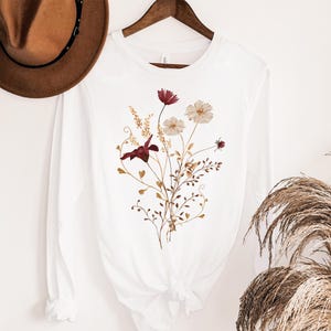 Op de afbeelding: Wit shirt met lange mouwen met een bloemenontwerp. Het ontwerp heeft bordeauxrode, cr&egrave;mekleurige en goudkleurige bloemen en bladeren. Het shirt hangt aan een houten hanger. Op de achtergrond is een bruine hoed te zien.