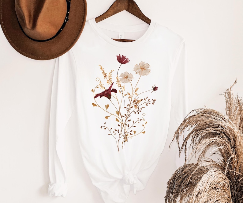 Op de afbeelding: Wit shirt met lange mouwen met een bloemenontwerp. Het ontwerp heeft bordeauxrode, cr&egrave;mekleurige en goudkleurige bloemen en bladeren. Het shirt hangt aan een houten hanger. Op de achtergrond is een bruine hoed te zien.