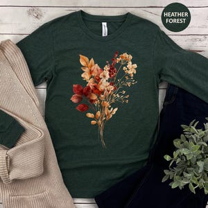 Pu&ograve; includere: Una t-shirt a maniche lunghe color verde bosco m&eacute;lange con una grafica centrale raffigurante un bouquet di fiori secchi nei toni dell'arancione, ruggine e crema. La maglia &egrave; abbinata a un cardigan in maglia beige, jeans blu scuro e una pianta.