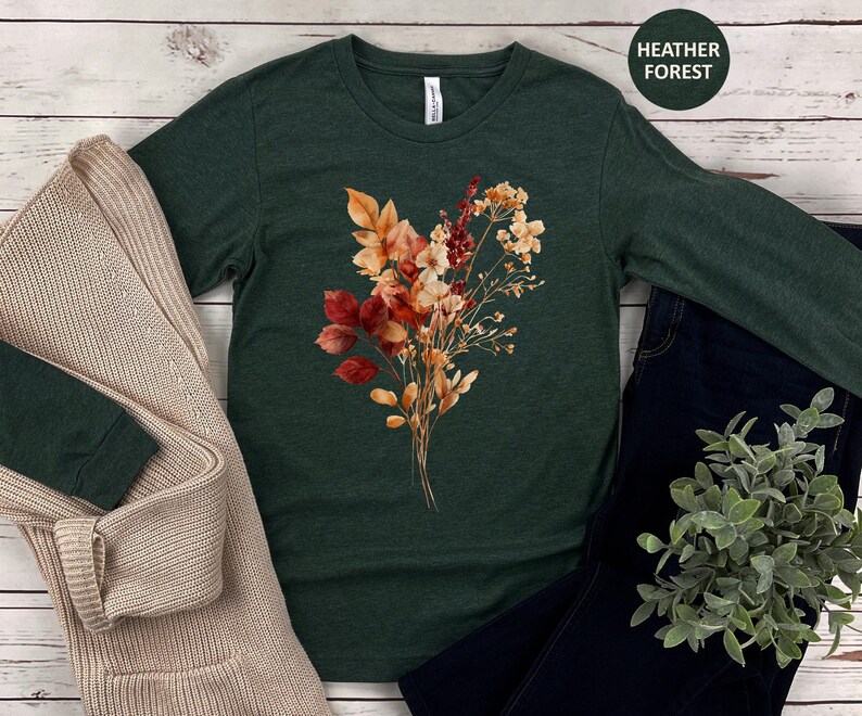 Pu&ograve; includere: Una t-shirt a maniche lunghe color verde bosco m&eacute;lange con una grafica centrale raffigurante un bouquet di fiori secchi nei toni dell'arancione, ruggine e crema. La maglia &egrave; abbinata a un cardigan in maglia beige, jeans blu scuro e una pianta.