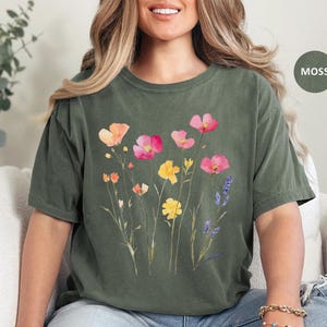 K&ouml;nnte beinhalten: Ein moosgr&uuml;nes T-Shirt mit einem Blumendruck mit rosa, gelben und orangefarbenen Blumen. Die Blumen sind im Aquarell-Stil gehalten und haben eine Vintage-&Auml;sthetik.