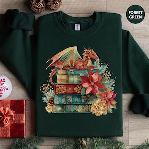 May include: Forest green sweatshirt featuring a watercolor illustration of a red dragon resting on a stack of books. The books are adorned with floral accents in red, yellow, and green. The sweatshirt has long sleeves and a crew neck.