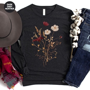 Op de afbeelding: Donkergrijze heather lange mouwen shirt met een bloemenontwerp in wit, rood en goud. Het shirt wordt gecombineerd met een blauwe jeans, een grijze hoed en een rode en teal sjaal.