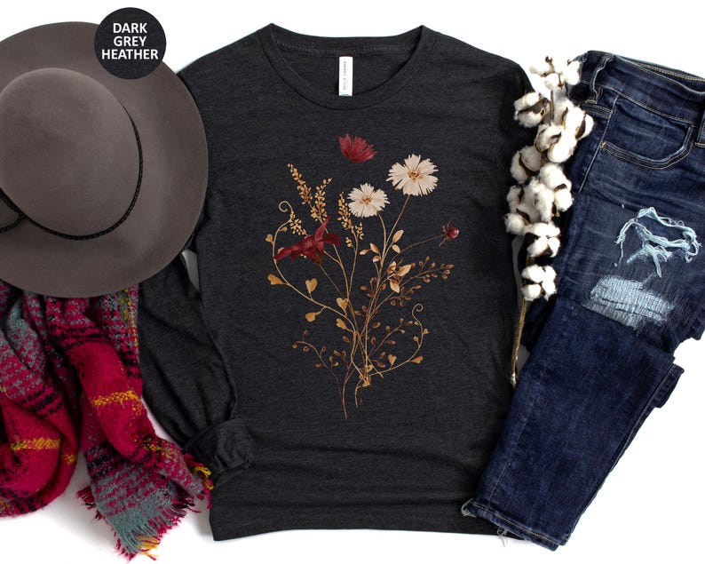 Op de afbeelding: Donkergrijze heather lange mouwen shirt met een bloemenontwerp in wit, rood en goud. Het shirt wordt gecombineerd met een blauwe jeans, een grijze hoed en een rode en teal sjaal.
