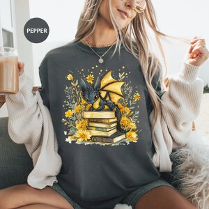 Puede incluir: Sudadera gris oscuro con una ilustraci&oacute;n fant&aacute;stica de un drag&oacute;n negro con alas amarillas sobre una pila de libros. El dise&ntilde;o incluye flores y follaje amarillos. La palabra "PEPPER" est&aacute; impresa en la parte superior izquierda.