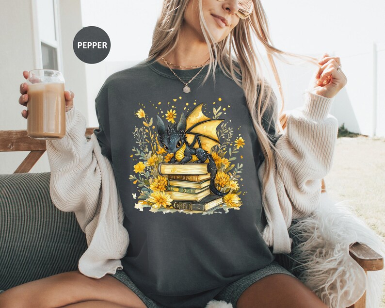 Puede incluir: Sudadera gris oscuro con una ilustraci&oacute;n fant&aacute;stica de un drag&oacute;n negro con alas amarillas sobre una pila de libros. El dise&ntilde;o incluye flores y follaje amarillos. La palabra "PEPPER" est&aacute; impresa en la parte superior izquierda.