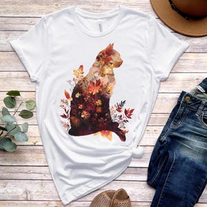 May include: White t-shirt featuring a cat silhouette filled with autumn leaves in shades of orange, red, and brown. The cat design is centered on the shirt. The shirt is paired with blue jeans and a brown hat.