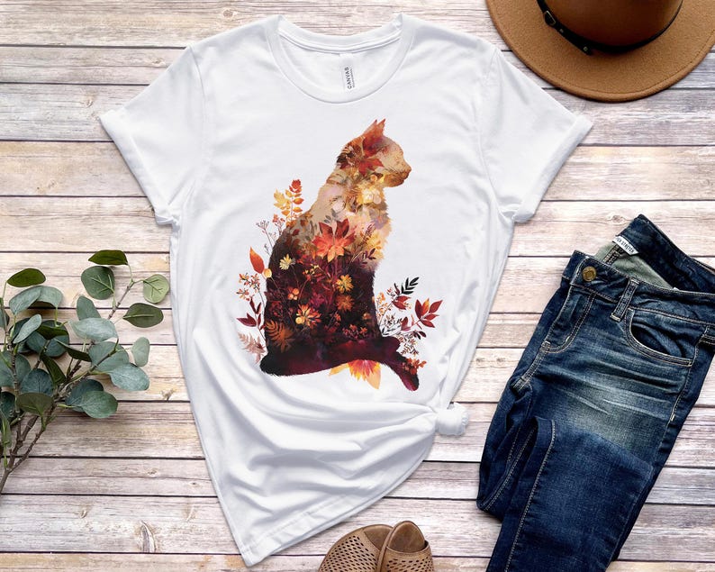May include: White t-shirt featuring a cat silhouette filled with autumn leaves in shades of orange, red, and brown. The cat design is centered on the shirt. The shirt is paired with blue jeans and a brown hat.