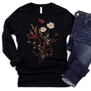 Op de afbeelding: Een zwart shirt met lange mouwen met een bloemenontwerp met rode, witte en goudkleurige bloemen en bladeren. Het shirt wordt gecombineerd met een donkerblauwe spijkerbroek. Het bloemenontwerp staat in het midden van de voorkant van het shirt.