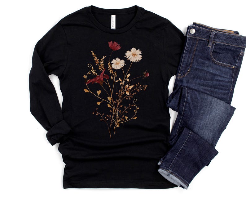 Op de afbeelding: Een zwart shirt met lange mouwen met een bloemenontwerp met rode, witte en goudkleurige bloemen en bladeren. Het shirt wordt gecombineerd met een donkerblauwe spijkerbroek. Het bloemenontwerp staat in het midden van de voorkant van het shirt.