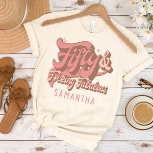 May include: A beige t-shirt with a pink graphic that says "Fifty & F*cking Fabulous" with the name Samantha underneath.