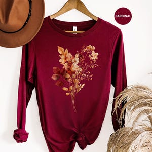 Pu&ograve; includere: Una maglia a maniche lunghe con girocollo color rosso cardinale, caratterizzata da una grafica centrale raffigurante un bouquet di fiori secchi con foglie e steli dai toni toni autunnali in sfumature di arancione, marrone e crema.