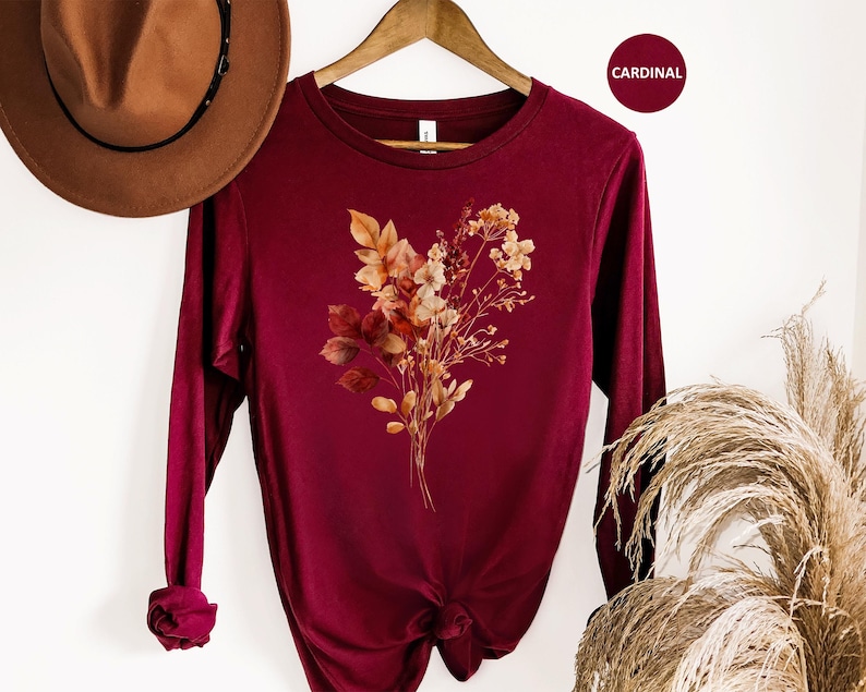 Pu&ograve; includere: Una maglia a maniche lunghe con girocollo color rosso cardinale, caratterizzata da una grafica centrale raffigurante un bouquet di fiori secchi con foglie e steli dai toni toni autunnali in sfumature di arancione, marrone e crema.