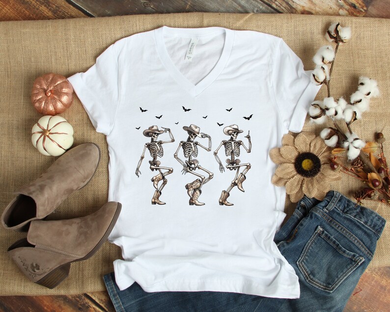 May include: White t-shirt with a graphic of three skeletons wearing cowboy hats and boots, dancing. Bats are flying above the skeletons.