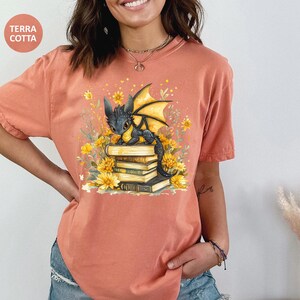 Puede incluir: Camiseta color terracota con una ilustraci&oacute;n de un drag&oacute;n negro con alas amarillas posado sobre una pila de libros, rodeado de flores amarillas. La camiseta tiene mangas cortas y un corte holgado. El texto "TERRA COTTA" est&aacute; en un c&iacute;rculo.