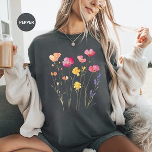 K&ouml;nnte beinhalten: Ein dunkelgraues Sweatshirt mit einem Blumendruck mit rosa, gelben und orangefarbenen Blumen. Die Blumen sind in einer Gruppe angeordnet und haben einen aquarellartigen Look. Der Text "PEPPER" ist in der oberen linken Ecke des Bildes sichtbar.