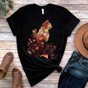 May include: Black t-shirt featuring a cat silhouette filled with autumn leaves in shades of red, orange, and yellow. The shirt is displayed with a brown hat and blue jeans on a wooden surface.