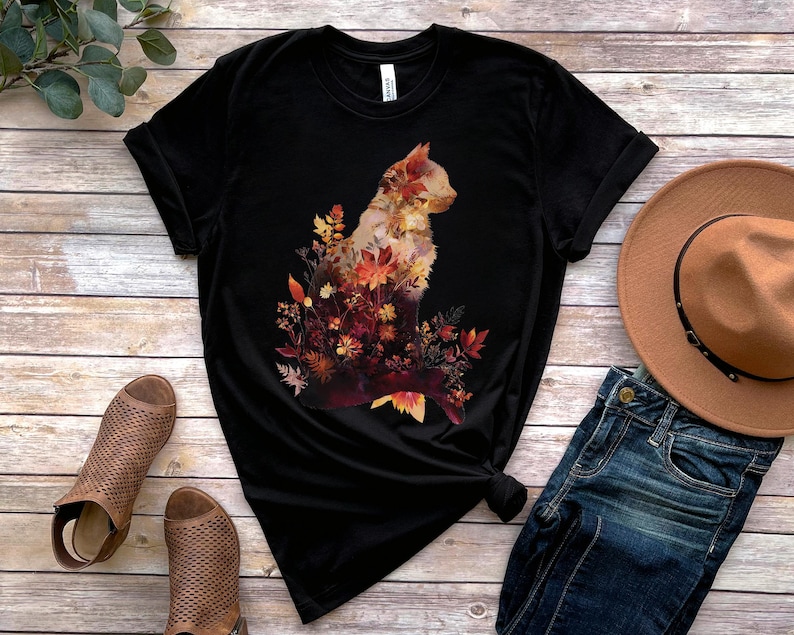 May include: Black t-shirt featuring a cat silhouette filled with autumn leaves in shades of red, orange, and yellow. The shirt is displayed with a brown hat and blue jeans on a wooden surface.