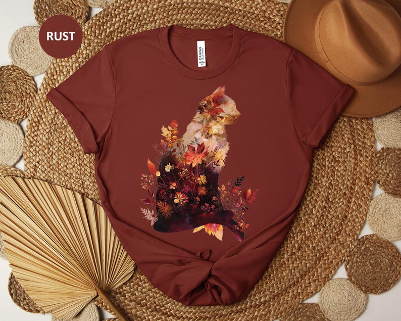 May include: Rust-colored t-shirt featuring a cat silhouette filled with autumn leaves in shades of red, orange, and yellow. The shirt has short sleeves and a crew neck. The word "RUST" is in a circle in the upper left corner.