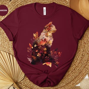 May include: Maroon t-shirt featuring a watercolor cat design filled with autumn leaves and flowers. The cat is silhouetted against the maroon fabric, with the word "Maroon" in a circle in the upper left corner.