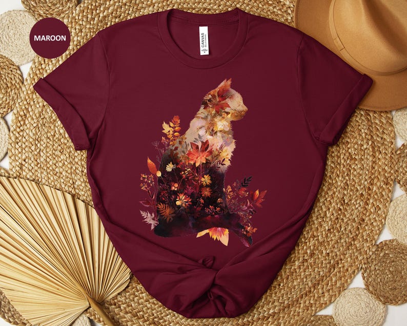May include: Maroon t-shirt featuring a watercolor cat design filled with autumn leaves and flowers. The cat is silhouetted against the maroon fabric, with the word "Maroon" in a circle in the upper left corner.