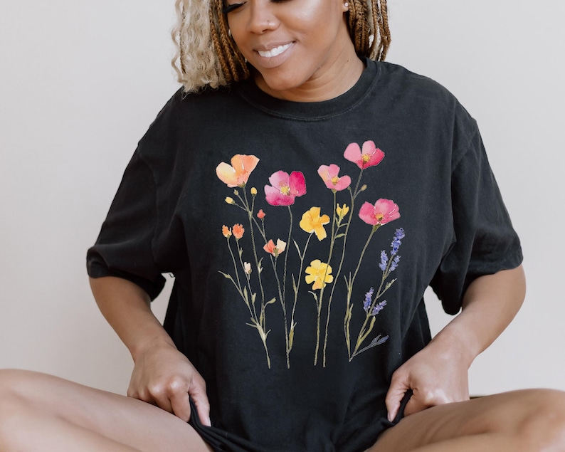 K&ouml;nnte beinhalten: Ein schwarzes T-Shirt mit einem Blumendruck, der rosa, orange und gelbe Blumen mit gr&uuml;nen Stielen und Bl&auml;ttern zeigt. Die Blumen sind in einer Gruppe auf der Vorderseite des Shirts angeordnet.