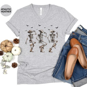 May include: A gray V-neck t-shirt with a graphic of three skeletons wearing cowboy hats and dancing. The skeletons are white with black details. The shirt has a distressed look and is made of a soft, comfortable material.