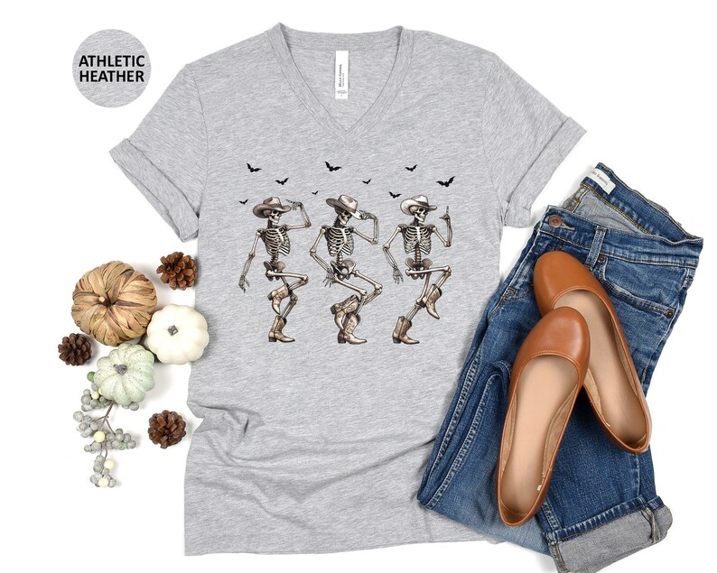 May include: A gray V-neck t-shirt with a graphic of three skeletons wearing cowboy hats and dancing. The skeletons are white with black details. The shirt has a distressed look and is made of a soft, comfortable material.