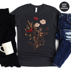 May include: A dark heather grey t-shirt featuring a floral design with dried flowers in shades of red, pink, and brown. The flowers are arranged in a bouquet-like shape and are surrounded by delicate vines.