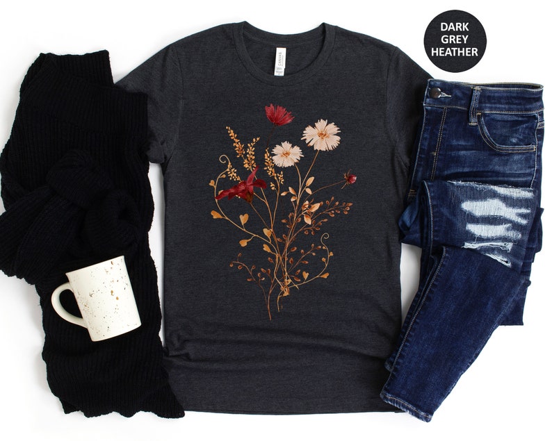 May include: A dark heather grey t-shirt featuring a floral design with dried flowers in shades of red, pink, and brown. The flowers are arranged in a bouquet-like shape and are surrounded by delicate vines.