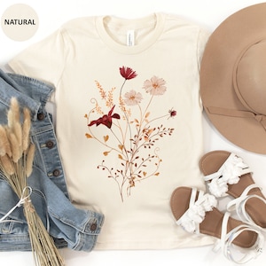 May include: A beige t-shirt with a floral print featuring red, pink, and brown flowers and leaves. The shirt is laid flat on a white surface with a denim jacket, a straw hat, and white sandals.