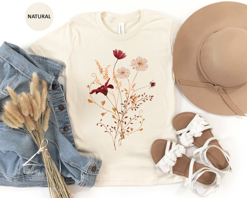 May include: A beige t-shirt with a floral print featuring red, pink, and brown flowers and leaves. The shirt is laid flat on a white surface with a denim jacket, a straw hat, and white sandals.