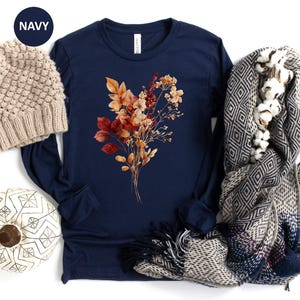 Pu&ograve; includere: Una maglia a maniche lunghe blu navy con una grafica centrale di fiori autunnali essiccati nei toni dell'arancione, rosso e marrone. &Egrave; accompagnata da un berretto in maglia beige, una zucca bianca con linee geometriche nere e una sciarpa grigia.