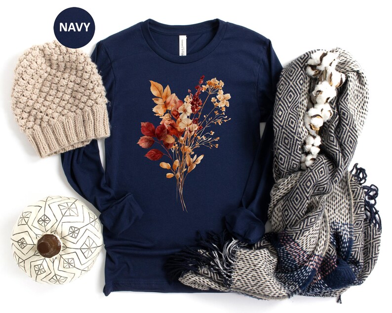 Pu&ograve; includere: Una maglia a maniche lunghe blu navy con una grafica centrale di fiori autunnali essiccati nei toni dell'arancione, rosso e marrone. &Egrave; accompagnata da un berretto in maglia beige, una zucca bianca con linee geometriche nere e una sciarpa grigia.
