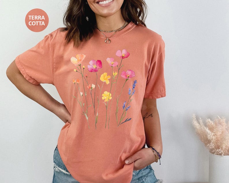 K&ouml;nnte beinhalten: Ein terracottafarbenes T-Shirt mit einem Blumendruck mit rosa, gelben und violetten Blumen. Die Blumen sind im Aquarell-Stil gehalten und in einer Gruppe auf der Vorderseite des Shirts angeordnet.