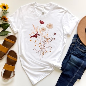 May include: White t-shirt with a floral print featuring burgundy, peach, and brown flowers. The shirt is styled with blue jeans, brown sandals, and a brown hat.