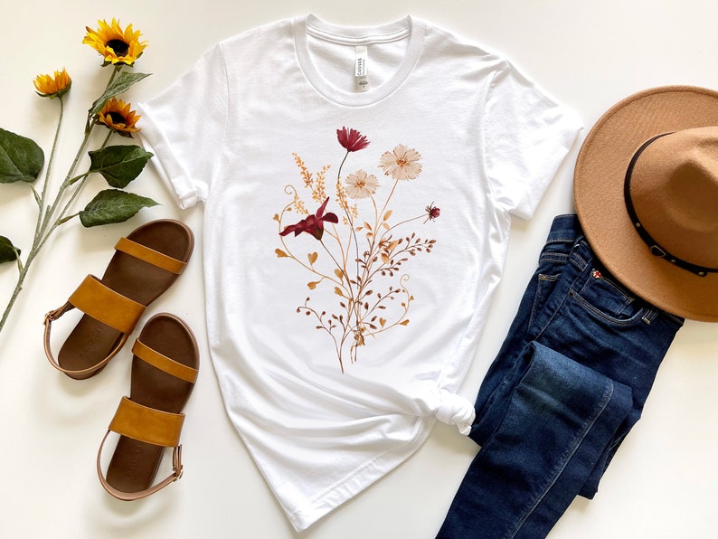 May include: White t-shirt with a floral print featuring burgundy, peach, and brown flowers. The shirt is styled with blue jeans, brown sandals, and a brown hat.