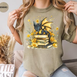 Puede incluir: Camiseta caqui con una ilustraci&oacute;n de un drag&oacute;n negro con alas amarillas sobre una pila de libros, rodeado de flores amarillas. El dise&ntilde;o est&aacute; impreso en una camiseta c&oacute;moda e informal.